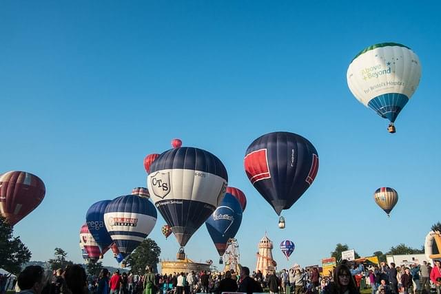 Multiple hot air balloons in a clear sky, a crowd of spectators gather in the foreground.
